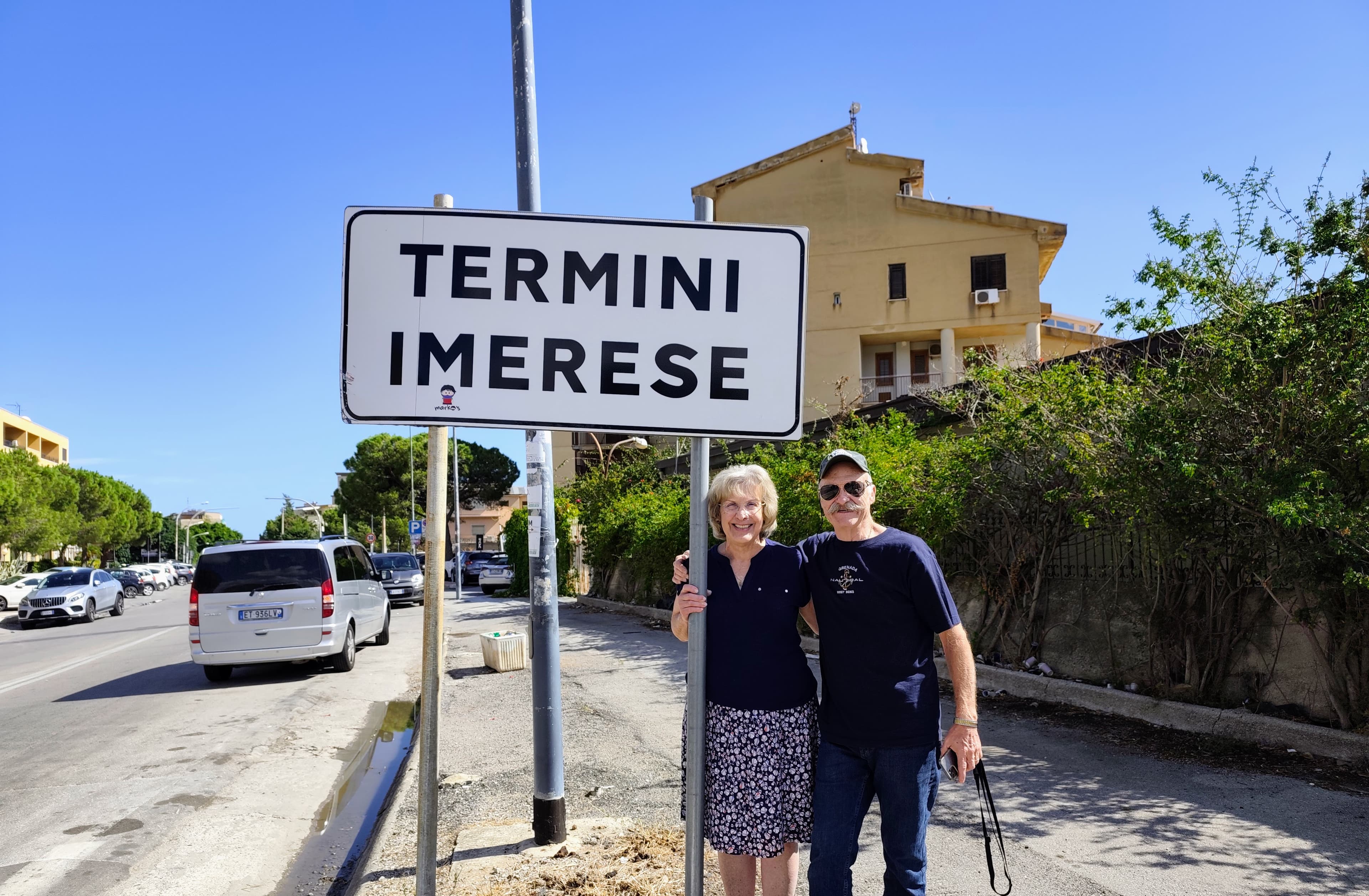 A smiling couple from US stands beneath the welcome sign for Termini Imerese, Sicily, during a heritage journey. The pair pose together on a sunny day beside the large town sign, with local streets, buildings, and greenery visible in the background under a clear blue sky.