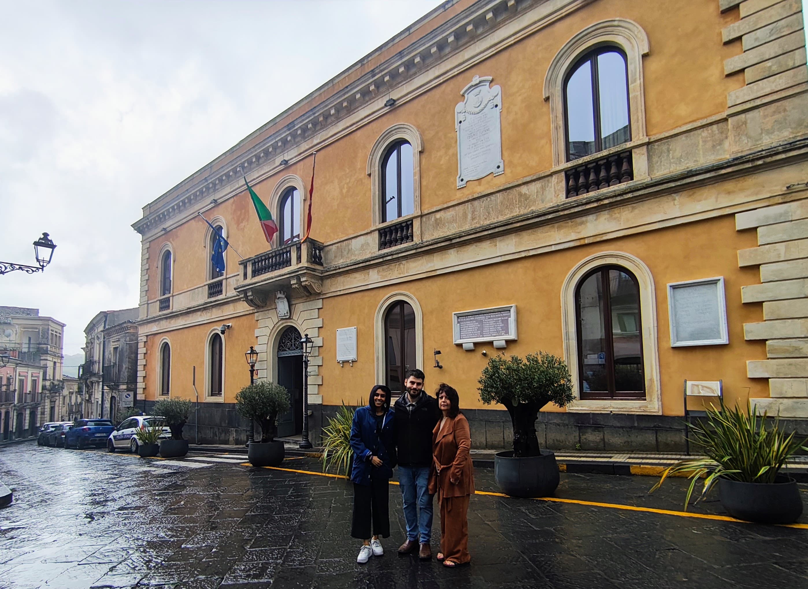 Mamatha and her family on a Sicily heritage tour standing outside a historic town hall building during a genealogy research visit. The group poses in front of the municipal building on a rainy day, where family records and ancestral documents may be preserved in Sicily.